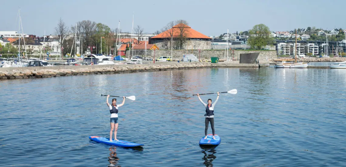 To personer på SUP-brett med Kristiansand festning i bakgrunnen.