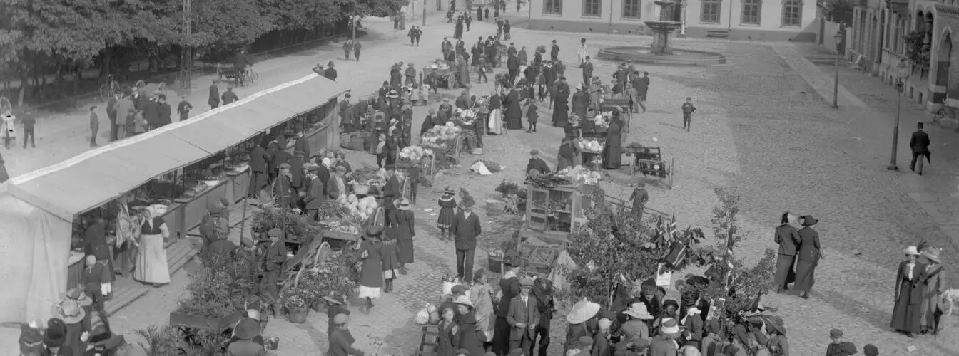 Bildet er i svart-hvitt og viser et mylder av mennesker som handler og prater sammen på torvet. Det er folk i alle aldere der. Ut fra klær og stil er bildet tatt cirka 1915.